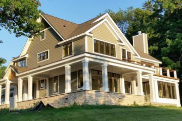 Yellow model home with wrap-around porch, white pillars, and brick on lower exposed foundation.