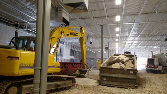 Excavator removing concrete slabs in an industrial building and loading it into a dump truck.