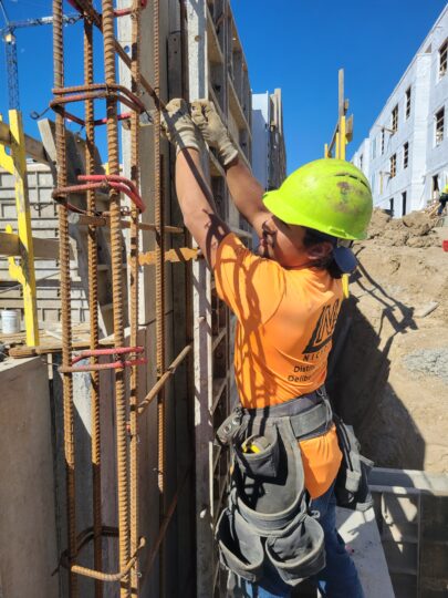 Construction worker in orange shirt and yellow hard hat with tool belt adjusting footings for concrete wall.